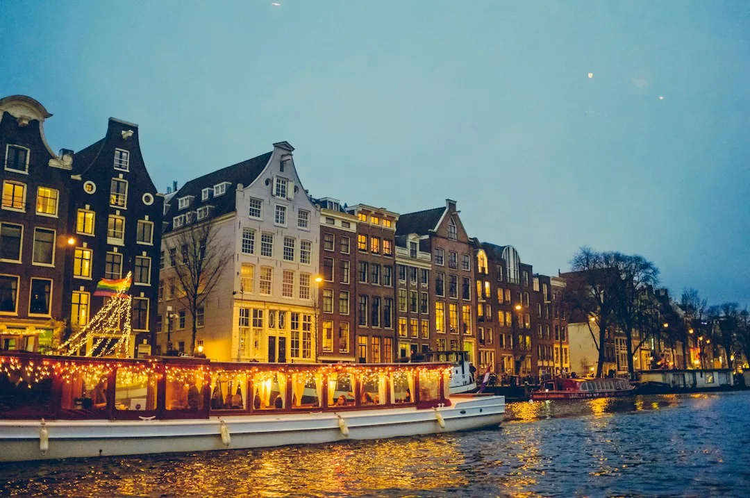 Canal de Ámsterdam al atardecer con hileras de casas históricas de fachadas tradicionales iluminadas, una de ellas decorada con luces navideñas, y barcas amarradas en el agua refleja.