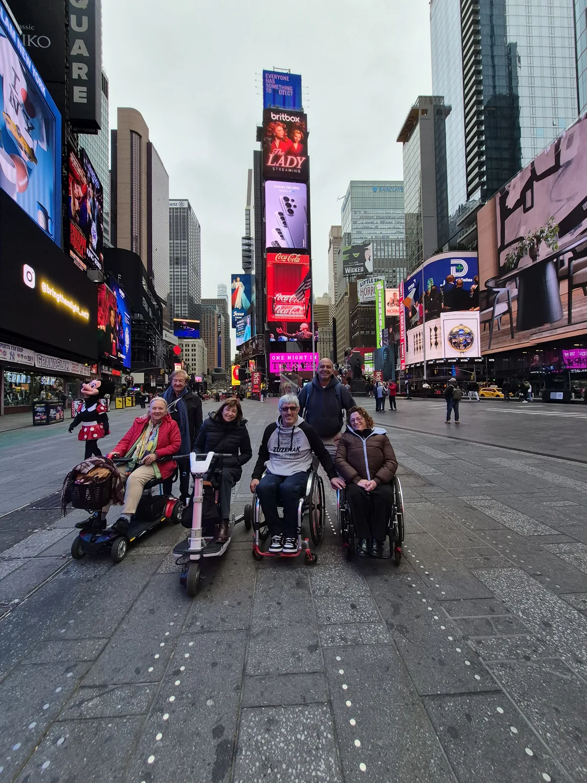 Times Square con grupo de personas en sillas de ruedas en primer plano sobre acera pavimentada, rodeados de edificios altos, pantallas digitales y señalética urbana.